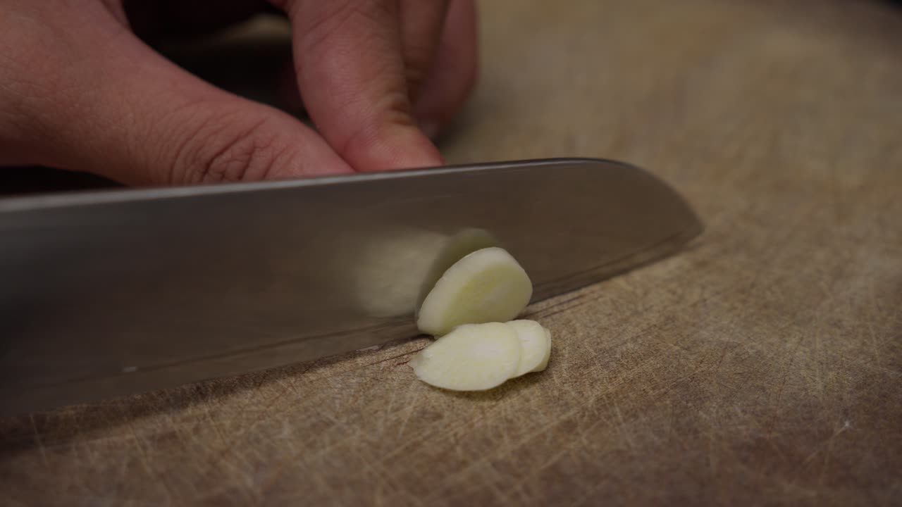 Close-up of hand slicing garlic on wooden cutting board.