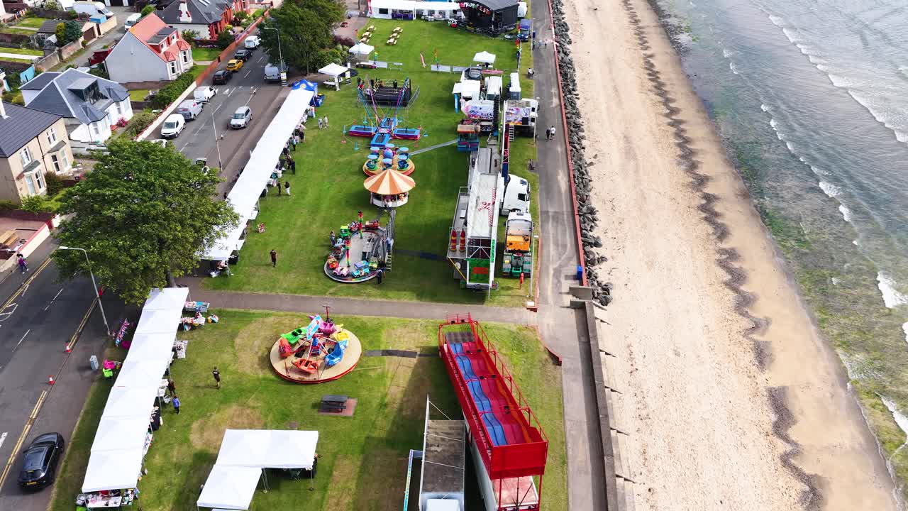 Drone glides above seaside fairground with rides, tents, and beach under bright daylight conditions