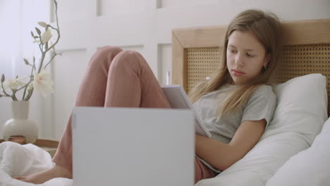 tired-and-bored-pupil-girl-is-lying-on-bed-with-exercise-book-and-listening-lesson-on-laptop-e-learning-for-school-children