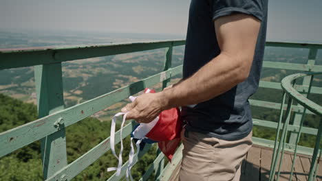 man reached top of lookout tower and taking a slovenian flag out of his backpack and tie it up on the fence