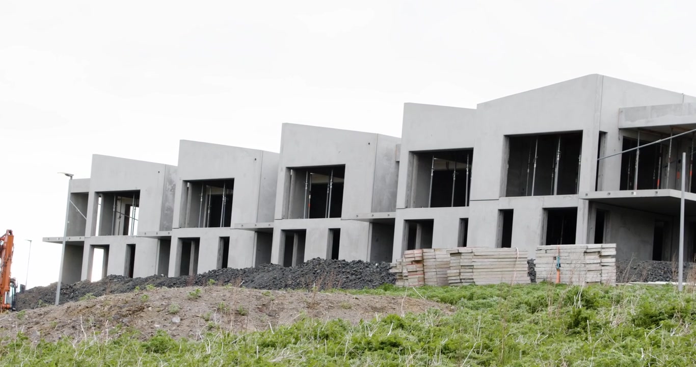 Een rij moderne, onafgewerkte betonnen huizen op een bouwplaats met hopen aarde en onkruid op de voorgrond onder een bewolkte hemel