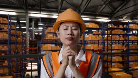 close up of asian female engineer with safety helmet prays for something while standing in the warehouse with shelves full of delivery goods