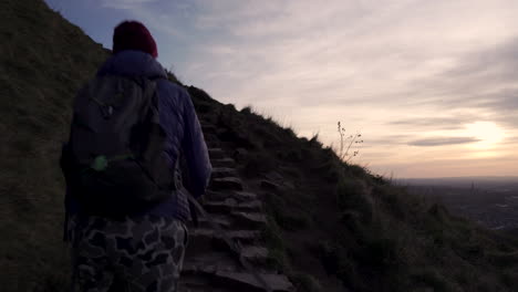 close up dolly shot of girl walking up the arthurs seat mountain on the hiking trail in evening during golden hour with city of edinburgh in the background down below