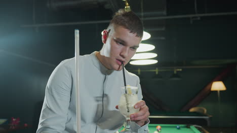 close-up of chap in white shirt holding cue stick, sipping lemon drink. camera zooms in, highlighting facial expression and moody ambiance of dimly lit pool hall with green table and overhead lighting