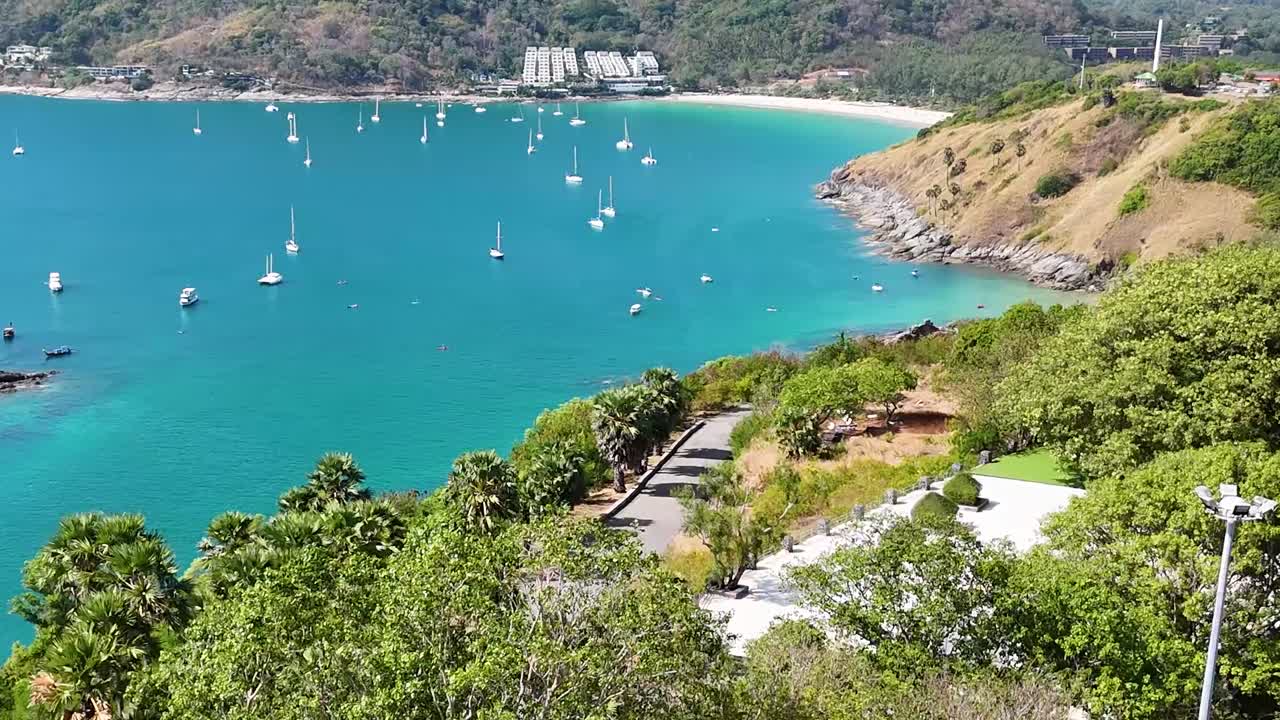 Aerial view of a vibrant bay with anchored boats, surrounded by lush greenery and a winding coastal road.