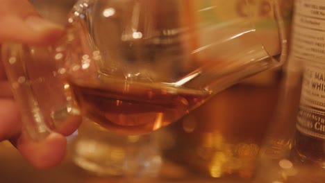 a close-up sequence of a hand gently swirling whisky in a traditional tasting glass, set against a warmly lit bar with bottles in the background