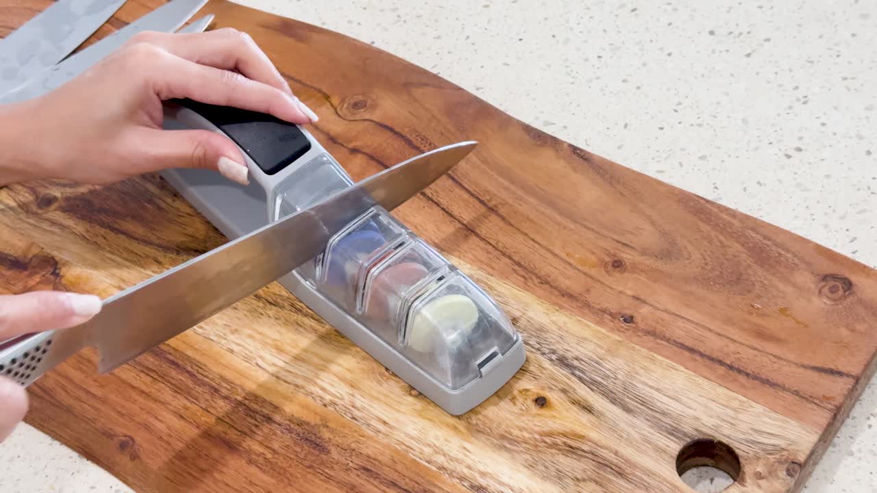 A person sharpens a kitchen knife using a manual sharpening stone on a wooden cutting board under bright, even lighting with steady overhead camera angles