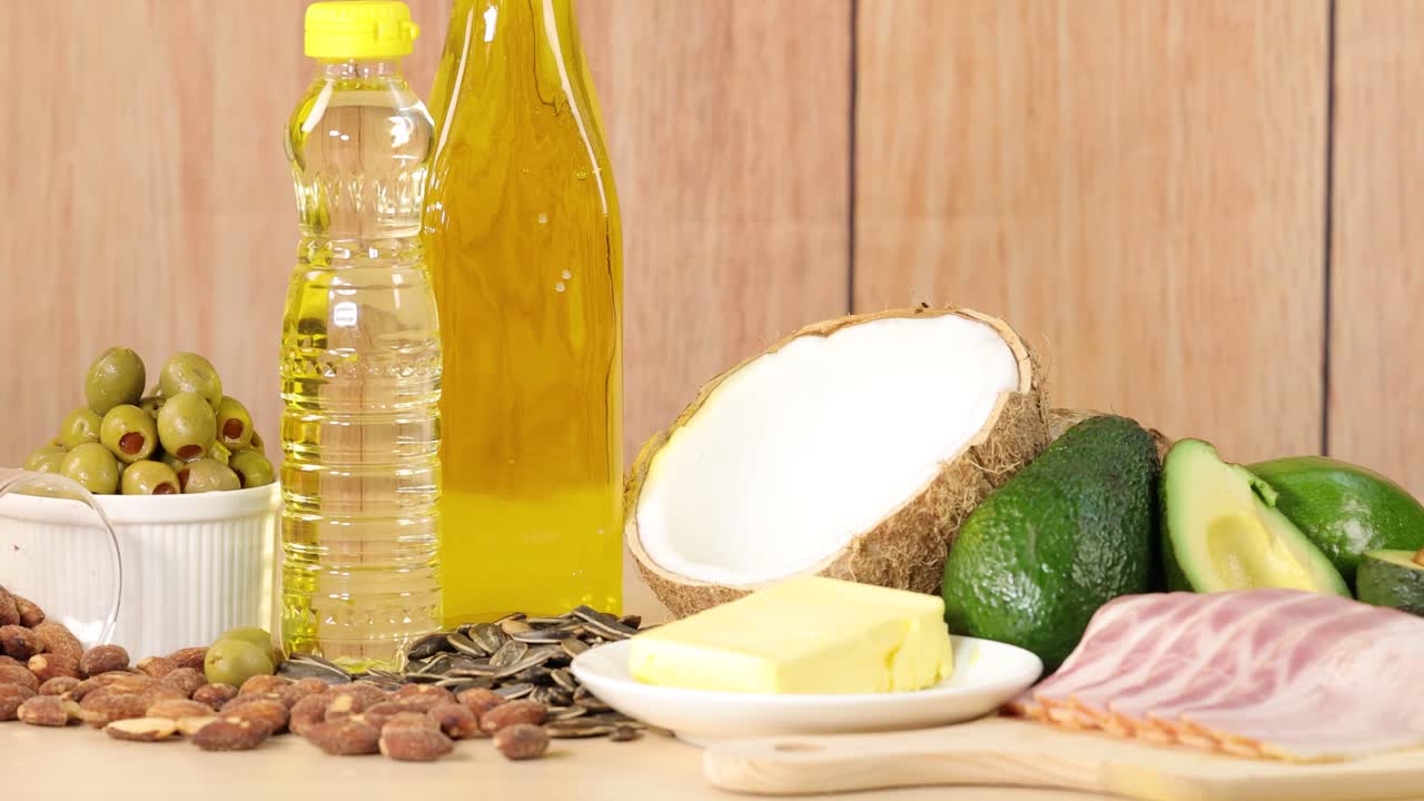 Various sources of dietary fat, including olive oil, coconut, butter, avocado, nuts, seeds, and olives, arranged on a wooden surface with soft, natural lighting and a slow camera pan