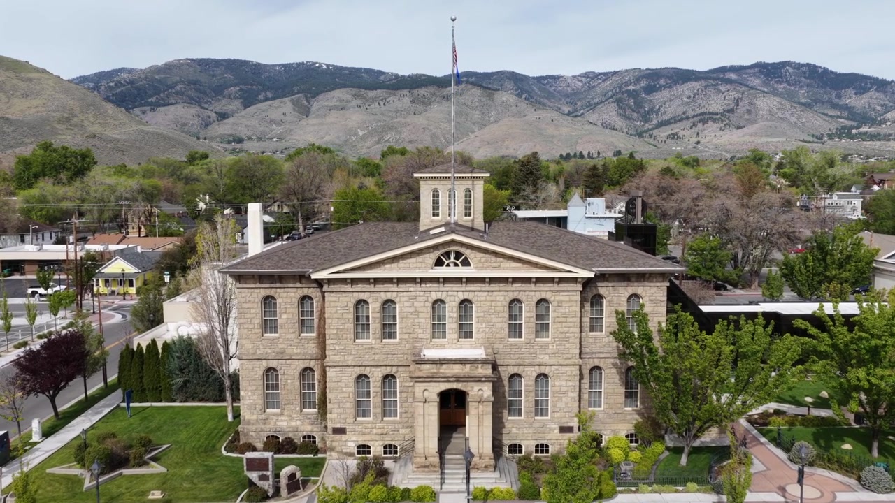 Historisk bygning for Nevada State Museum i Carson City med flagg som vaier over steinfasaden og kuppelen plassert blant grønne trær med ørkenfjell under klar himmel på dagtid. Luftfoto som nærmer seg