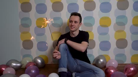 Young-Man-Sitting-with-a-Sparkler-Against-Colorful-Background,-Surrounded-by-Balloons-on-the-Ground