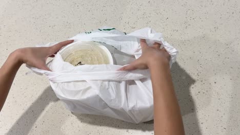 hands carefully unwrap a plastic bag to reveal a container of noodles on a light countertop