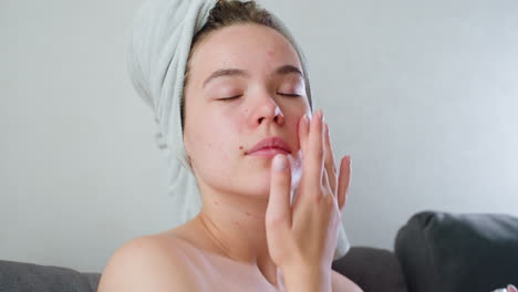 close up view of woman with acne-prone skin applying skincare cream on face using fingers while relaxing on couch, wrapped in towel with hair covered, enjoying gentle self care routine