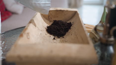 hand view of female plant lover wearing white gloves spreading black sand into wooden container on glass table beside potted branches for indoor gardening in cozy brick walled growing room