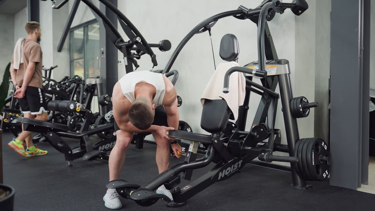 Weight lifter adjusts machine seat then sits gripping handle firmly, body engaging muscles under gym lighting as he positions for intense chest press workout on sleek equipment with dynamic motion