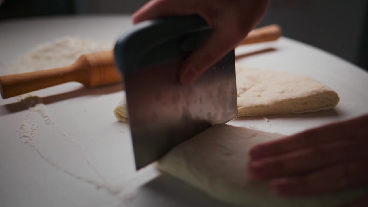 Una mano taglia l'impasto con un coltello su una superficie infarinata. Un mattarello riposa nelle vicinanze. La scena mostra un primo piano del processo di preparazione della pizza. Ideale per progetti culinari o legati al cibo.