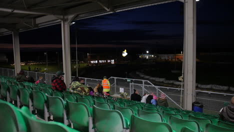 sparse spectators sit in green stadium seats under an open-air roof, overlooking a track at dusk. the dark sky and distant lights create a quiet, expectant atmosphere at the outdoor venue