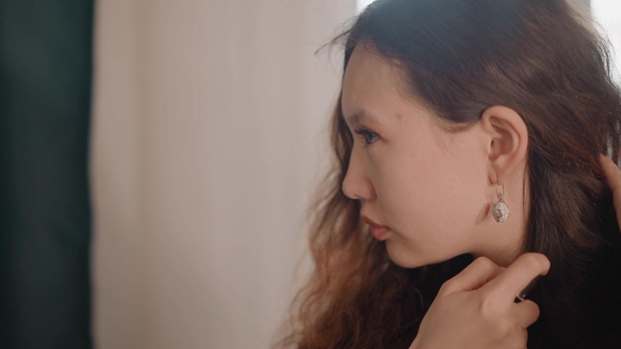Profile view of young woman securing dangling earring with fingers while tucking hair behind ear, set in soft window light with calm expression, natural skin tones, and elegant beauty routine mood