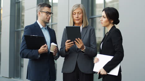 Mujer-De-Negocios-Madura-Leyendo-Un-Proyecto-De-Negocios-En-Una-Tableta-Mientras-Habla-Con-Sus-Colegas-En-La-Calle