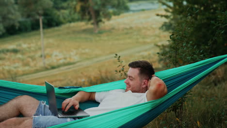 combining work and pleasure. caucasian man lies in a hammock and works on laptop. nature at backdrop.