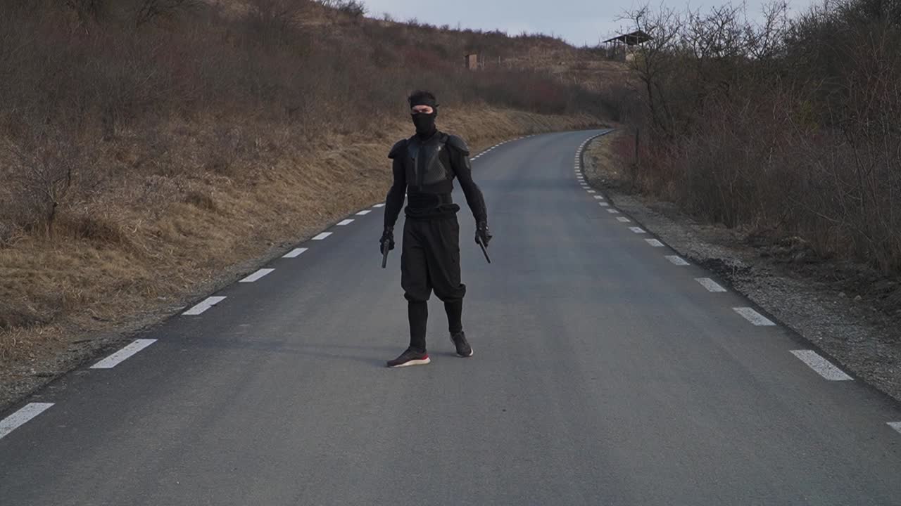 A lone ninja dressed in black tactical armor stands in the middle of road, arms extended, holding two pistols. A cinematic scene for action films, martial arts content, and storytelling visuals.