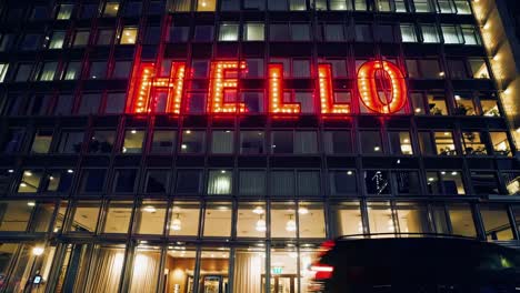 glowing red hello sign illuminating modern glass building facade during nighttime, showcasing urban cityscape with passing car and varying perspectives
