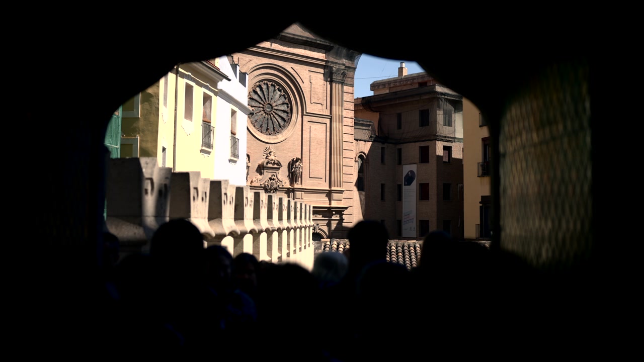 Strada storica cittadina illuminata dal sole appare attraverso un arco scuro, con facciata di chiesa ornata, rosone circolare, facciate color pastello, tetti in tegole e pedoni in silhouette lungo il bordo inferiore