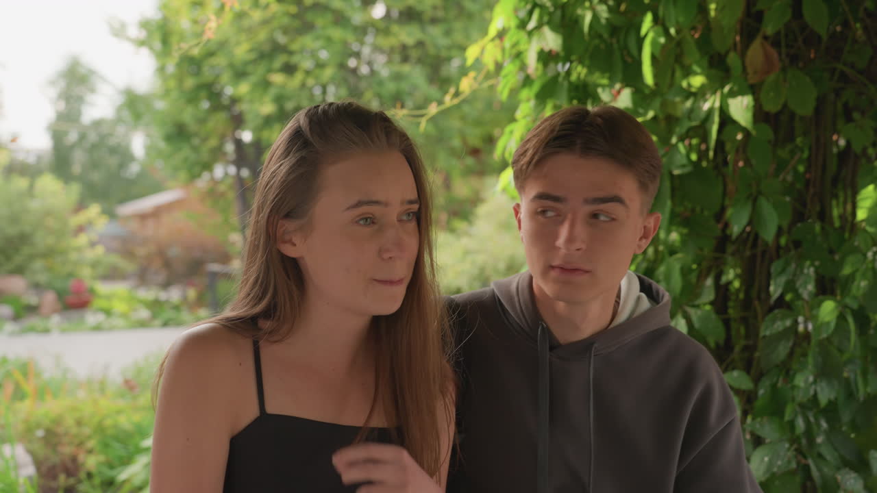 Una coppia di adolescenti sotto una pergola di vite si scambia sguardi timidi e sorrisi silenziosi, luce pomeridiana soffusa, passerella suburbana incorniciata da verde fogliame, felpa casual e abito nero, momento di amicizia intima, delicato