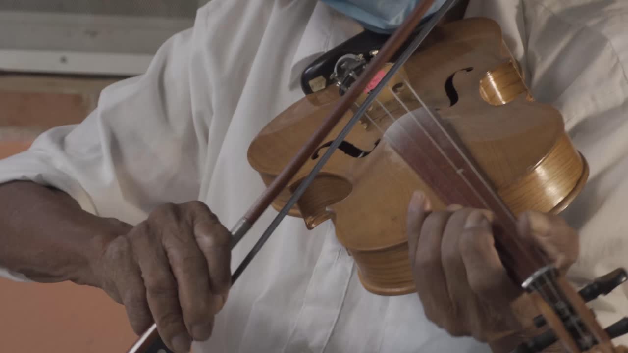 captura de pantalla de un músico de edad hispana con un sombrero y una máscara tocando el violín en la calle