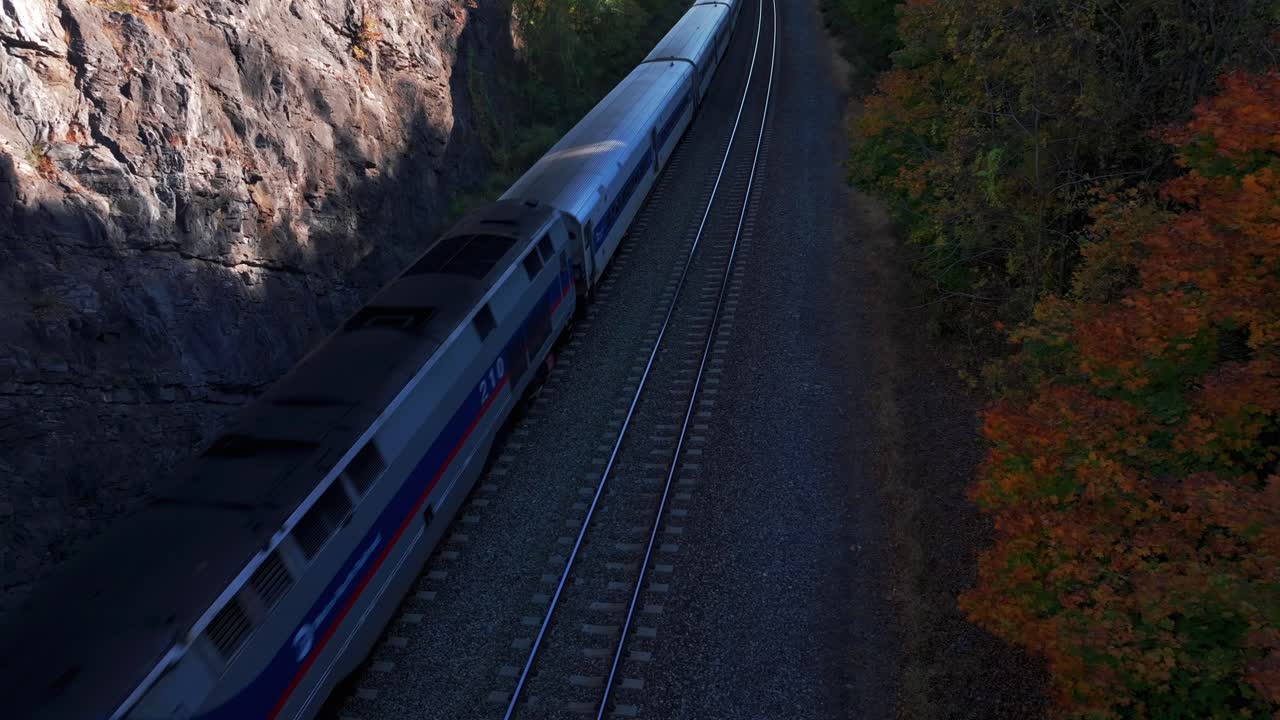 An aerial view of train tracks in a deep ravine upstate on a sunny day in autumn. The camera is stationary, tilted downward as an MTA train passes between a rock cliff and colorful trees.