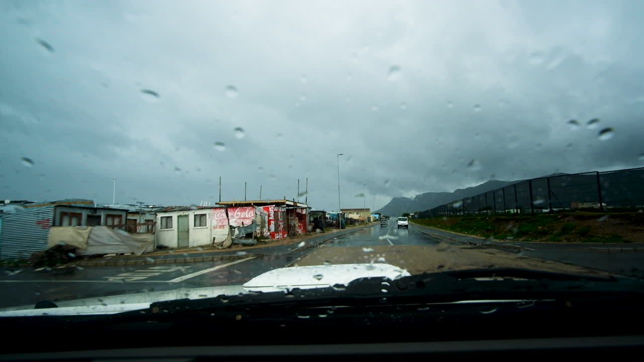 Car drives in rain through township with informal houses; apartheid concept