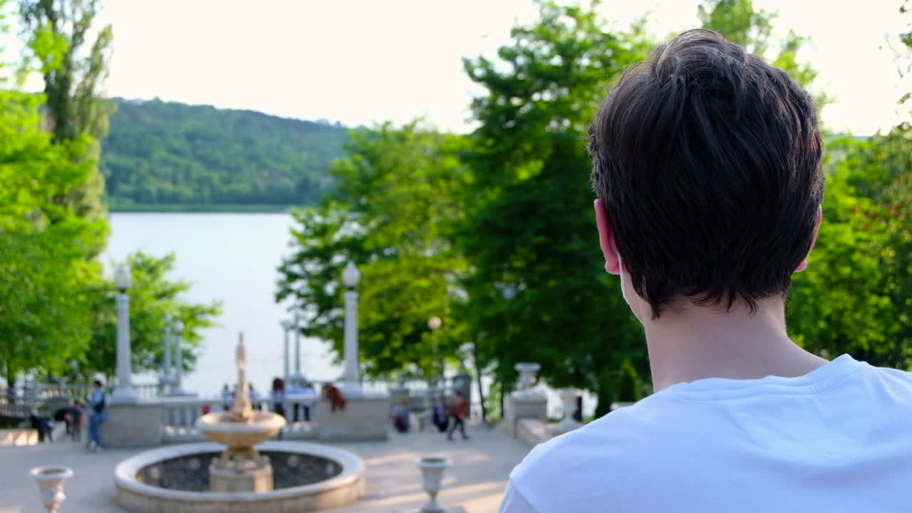 Young man in white t-shirt and sunglasses taking off a protective medical mask from the face while looking at the lake and walking people in park. Sunny day. Rear view. Corona Virus idea