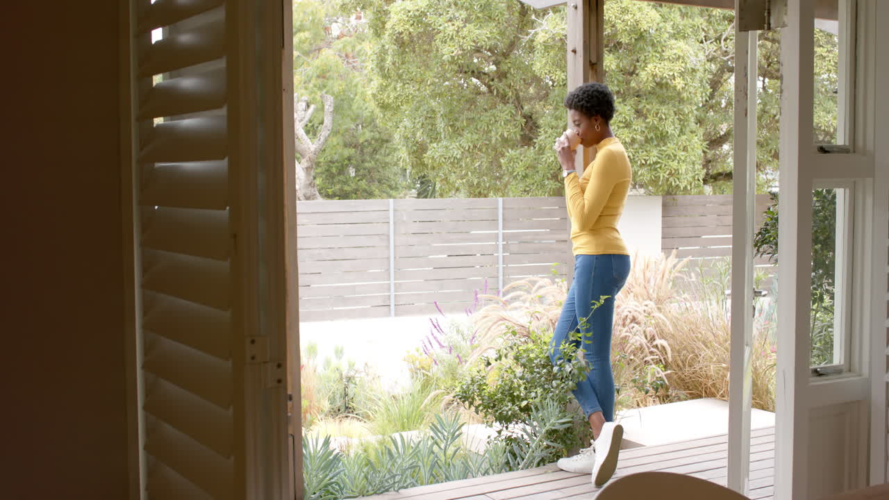 African american woman standing on porch, drinking coffee and enjoying garden view at home