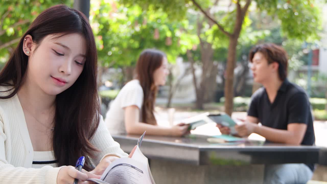 一位年輕亞洲女性在戶外校園中學習時，正在閱讀教科書並微笑