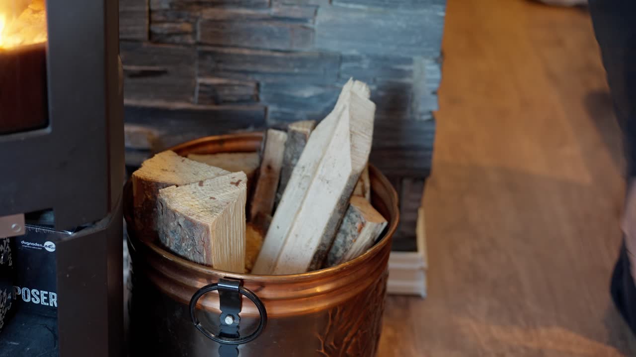 Man fills copper colored bucket near wood burning stove with firewood during winter.