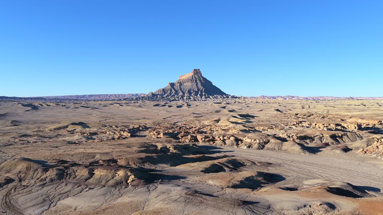 Un ampio scatto aereo con drone cattura Factory Butte, un elemento geologico prominente nello Utah. L'isolato butte si erge su un vasto deserto brullo e su un terreno di badlands grigio sotto un cielo azzurro.