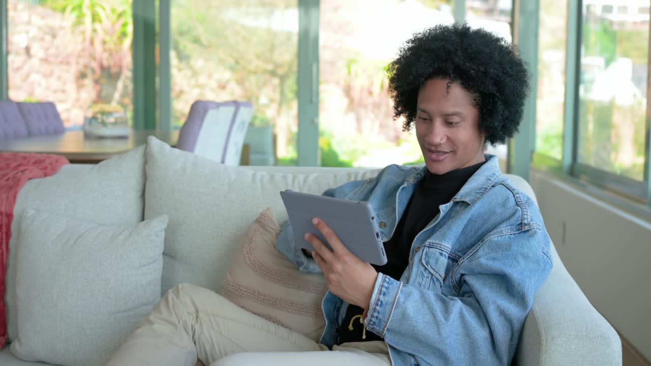 Dopo essersi sistemato sul divano, un uomo vestito di denim scorre su un tablet, allunga la mano verso una tazza e sorseggia il caffè a casa