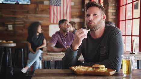 man watching game on screen in sports bar eating burger and fries and drinking beer