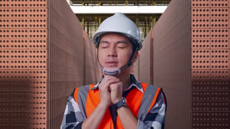 close up of asian male engineer with safety helmet pray for something while standing with red brick packed in stacks are stored