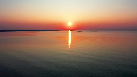 Vista-Aérea-Por-Drones-Del-Hermoso-Amanecer-En-El-Océano-Con-Un-Sol-Brillante-Que-Proyecta-Coloridos-Reflejos-Rojos,-Naranjas,-Morados-Y-Amarillos-Sobre-Las-Tranquilas-Olas-Del-Mar-Con-Un-Hermoso-Cielo-Y-Agua