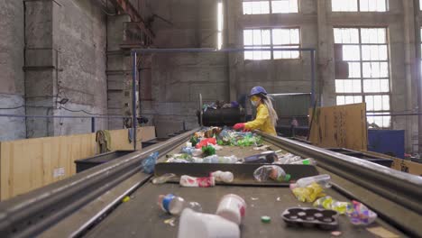 woman-volunteer in yellow and transparent protecting glasses, hard hat and mask sorting used plastic bottles at recycling plant. separate bottles on the line, removing tops and squeeze them.low angle footage from the conveyor