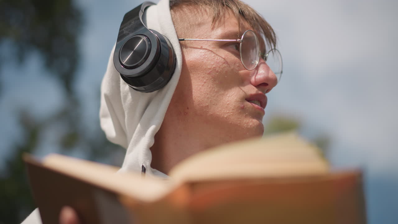 헤드폰을 끼고 야외에서 책을 읽고 있는 젊은 백인 남성, 후드티, 둥근 안경, 페이지를 넘기는 모습, 흐린 하늘과 부드러운 햇빛, 생각에 잠긴 표정, 공부하다가 딴생각에 잠기는 모습, 학생을 보여주는 프레임