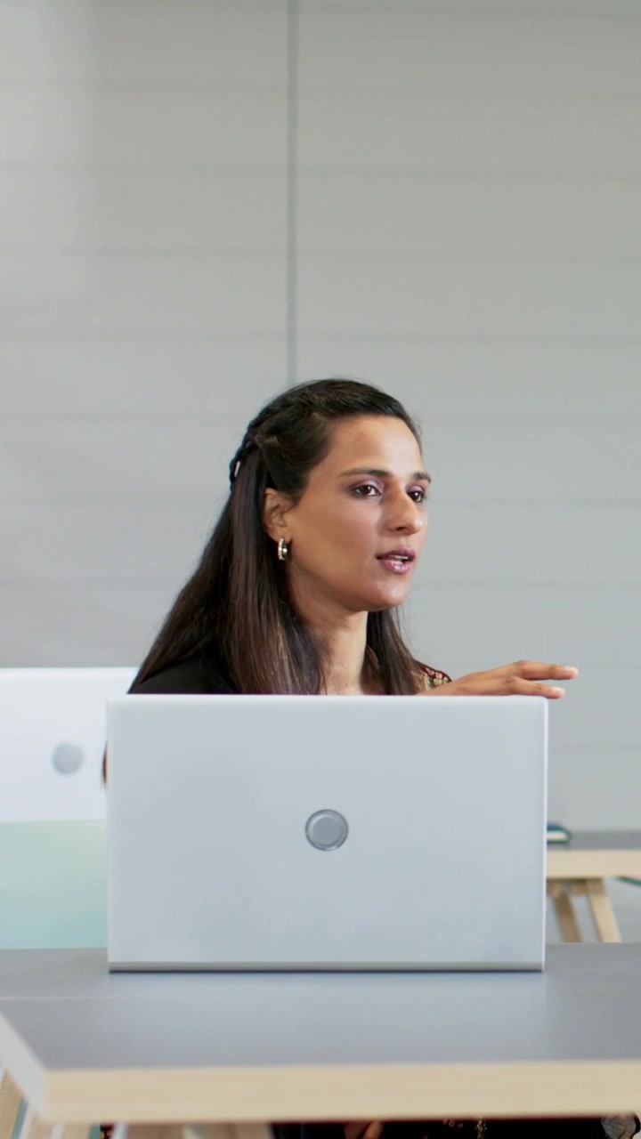 Video vertical: Mujer sonriente con top oscuro mirando la pantalla y hablando a la cámara en un escritorio con un portátil.