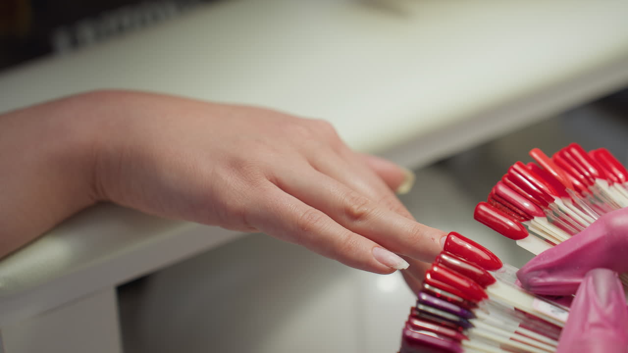 Closeup of nail technician placing red nail polish sample tip on customer finger for comparison during color selection. Various red and glittery shades displayed for client decision at nail salon