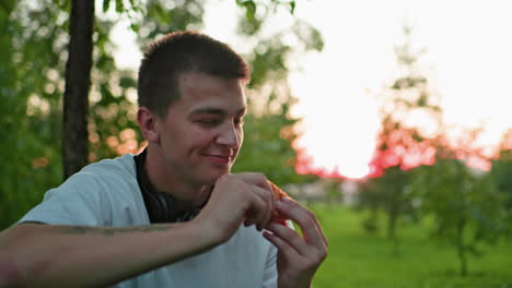 man with tattooed hand smiling while making a toast with bread, with a partial view of another person holding bread in the background, sunlight filters through trees