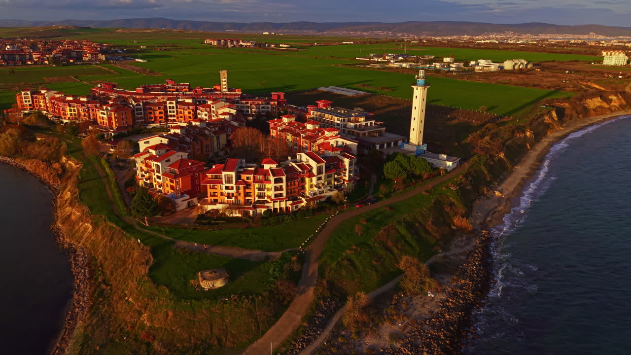 Villaggio costiero con edifici luminosi e uno storico faro che si affaccia sui campi e sull'oceano circostanti al tramonto