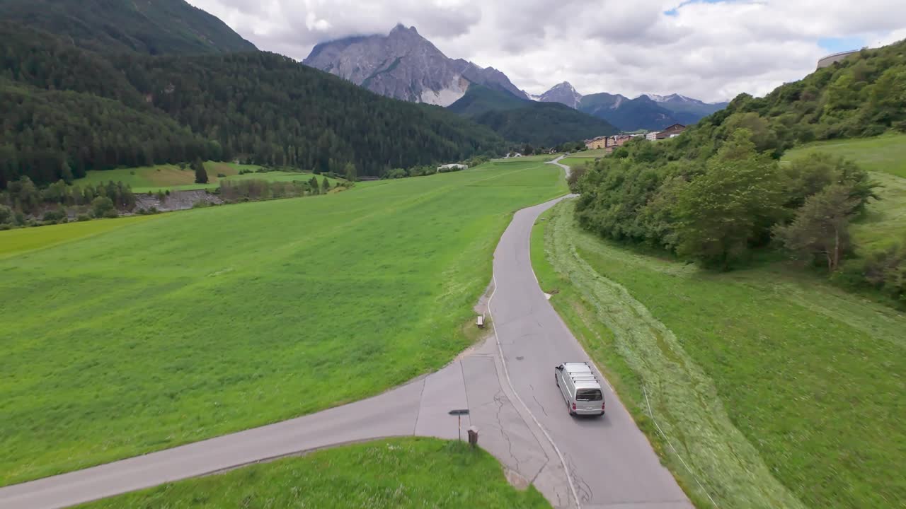 Drone tracking shot of silver transporter van car on Swiss road between idyllic fields and alp mountains. Following shot