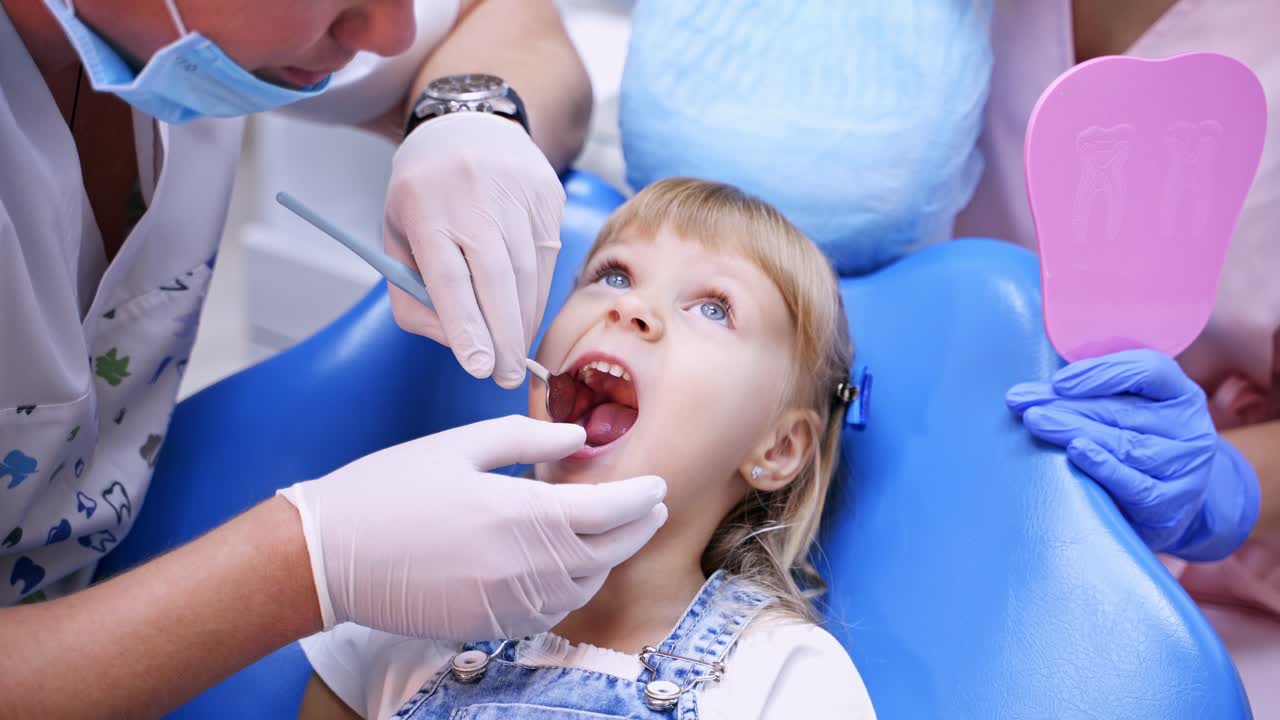 Little child in stomatology chair reseives treatment. Doctor is checking girl's teeth with dentist mirror - close up video.