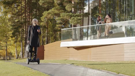 giovane uomo d'affari elegante che guida uno scooter mentre utilizza lo smartphone sulla strada nel parco 1