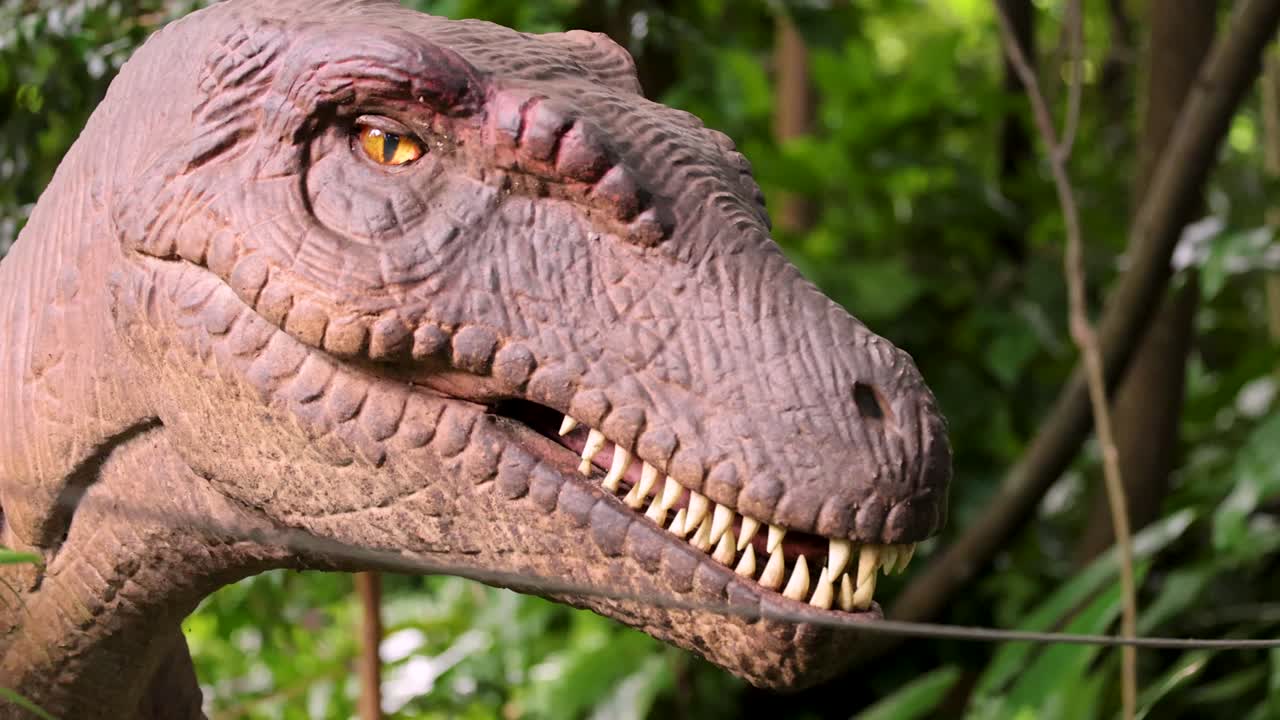 A realistic animatronic Velociraptor head turns and blinks in a sunlit, green outdoor park environment, with close-up framing and natural lighting