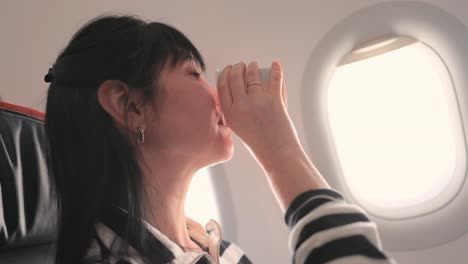 Woman-sitting-inside-airplane-and-looking-at-window-and-drinks-coffee.